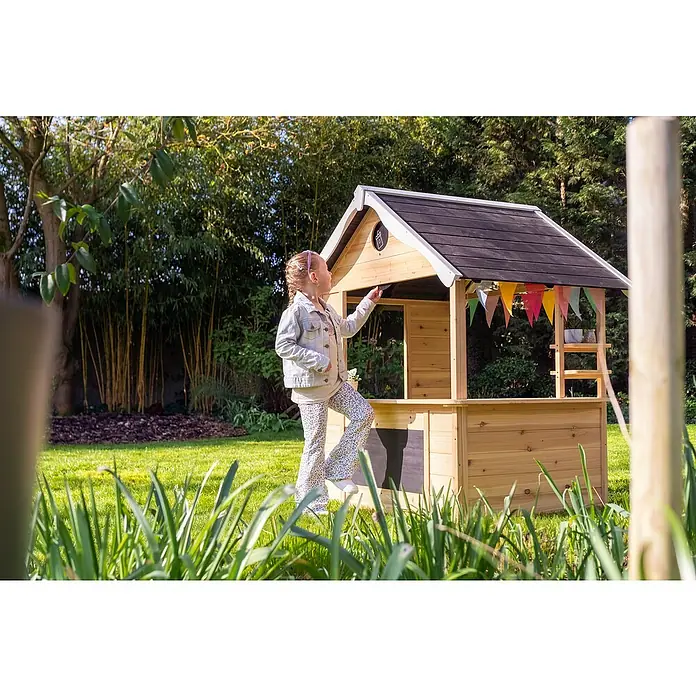 Exit Toys Spielhaus Hika - L x B x H: 140 x 111 x 155 cm, Holz, Natur - Holzspielhaus mit dunklem Dach und bunter Wimpelkette, ein Kind spielt damit im sonnigen Garten.