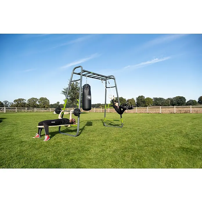 Exit Toys Klettergerüst GetSet Monkeybar MB230 - 442 x 125 x 248 cm, Stahl, Grau - Zwei Personen nutzen ein grau-grünes Outdoor-Fitnessgerät. Eine drückt Gewichte, die andere hängt an Turnringen. Ein schwarzer Boxsack ist ebenfalls angebracht.
