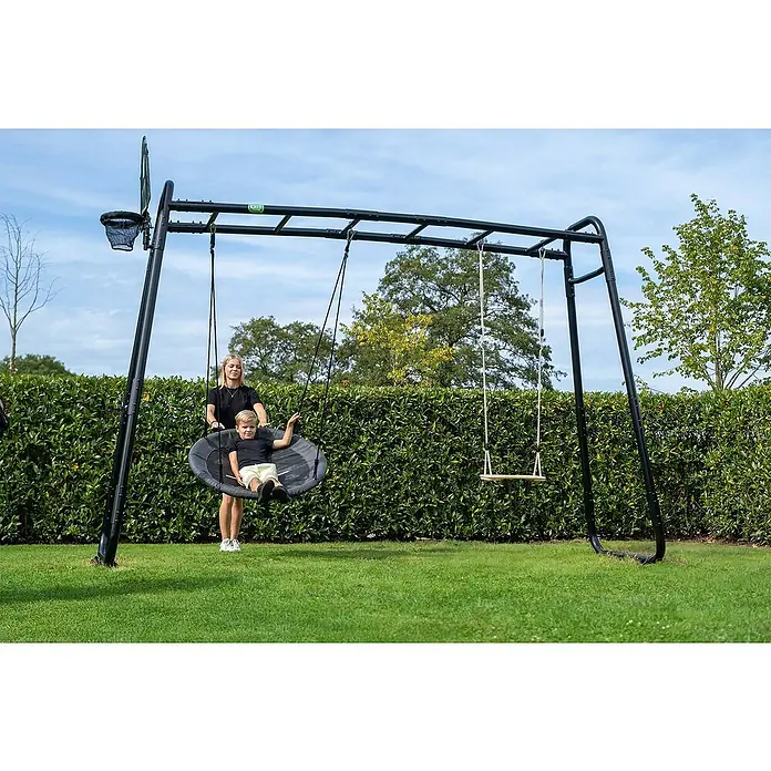 Exit Toys Nestschaukel - Durchmesser: 100 cm, Metall, Schwarz/Grau - Schwarzes Metall-Schaukelgestell mit Korbschaukel, Brettschaukel und Basketballkorb, von Frau und Kind im Garten genutzt.