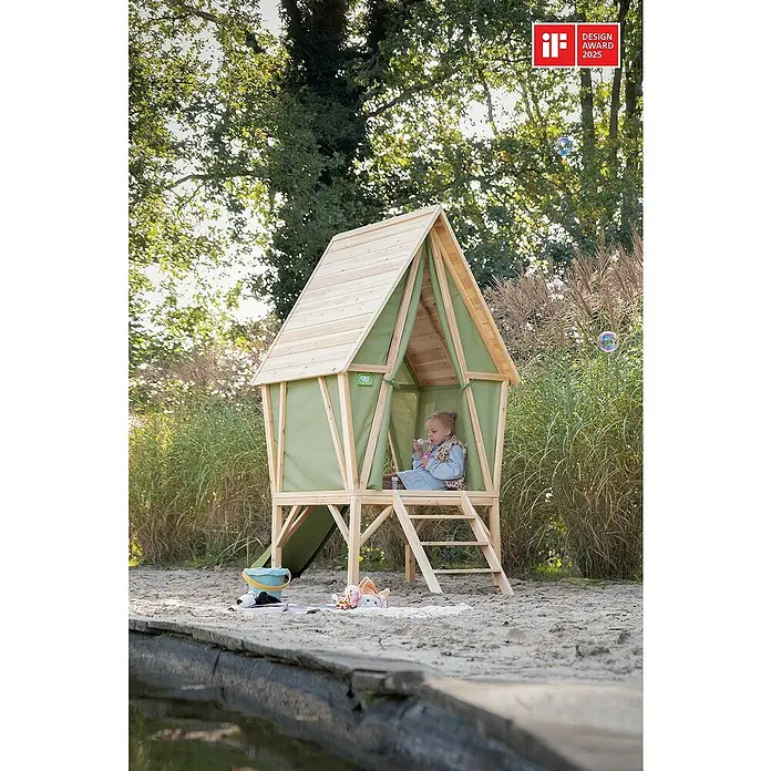 Exit Toys Spielhaus Yuki 300 - L x B x H: 226 x 124 x 219 cm, Holz, Natur/Grün - Erhöhtes Holz-Spielhaus mit grünen Stoffwänden auf Sand, ein Kind bläst darin Seifenblasen.