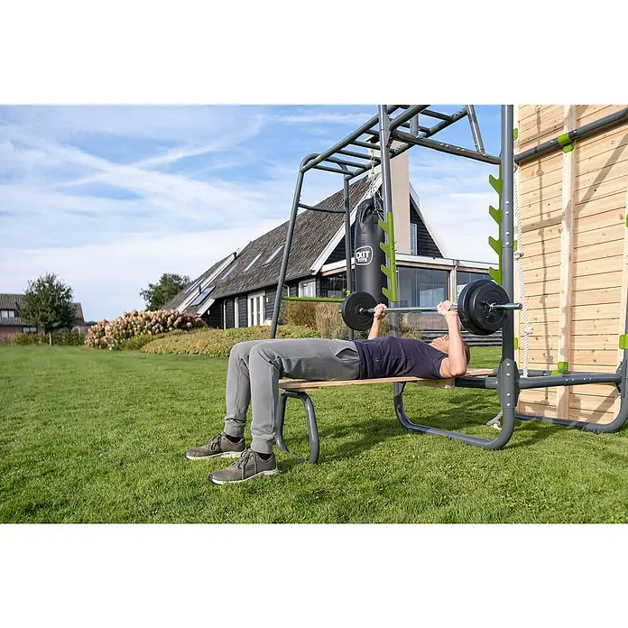 Exit Toys Klettergerüst Trainingsbank GetSet - Passend für: GetSet Monkeybars & Powerstations MB200/MB 300 & PS500/PS600, 115 x 30 x 41 cm, Natur - Mann beim Bankdrücken an einer grauen und hölzernen Outdoor-Fitnessstation mit Boxsack und Kletterwand.