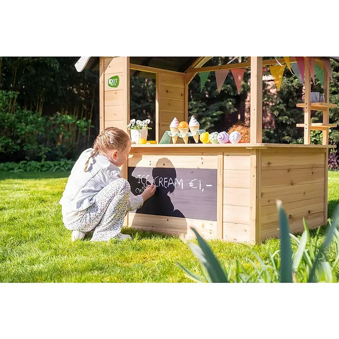 Exit Toys Spielhaus Hika - L x B x H: 140 x 111 x 155 cm, Holz, Natur - Ein Kind schreibt auf die Tafel eines hölzernen Spielzeugladens im Garten.