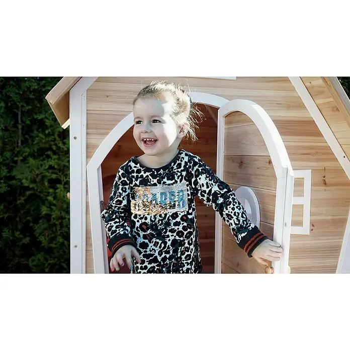 Exit Toys Spielhaus Fantasia 100 - L x B x H: 128 x 108 x 171 cm, Holz, Natur - Ein glückliches Kind spielt in einem Spielhaus aus Naturholz mit weißem Besatz, das seinen Gebrauch im Freien zeigt.