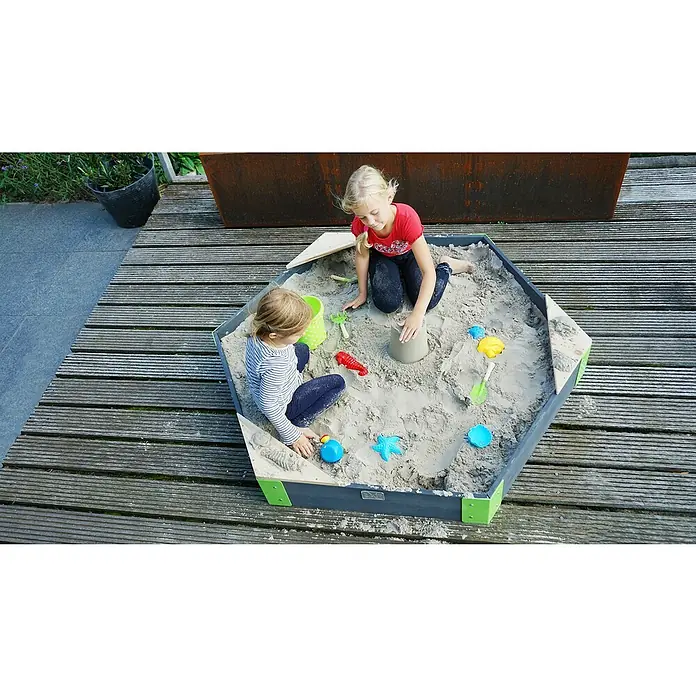 Exit Toys Sandkasten Aksent Sechseck - 160 x 140 x 21 cm, Holz, Grau - Zwei Mädchen spielen in einem sechseckigen Sandkasten mit grauen Seiten und grünen Akzenten, gefüllt mit Sand und Spielzeug, auf Holzdeck.