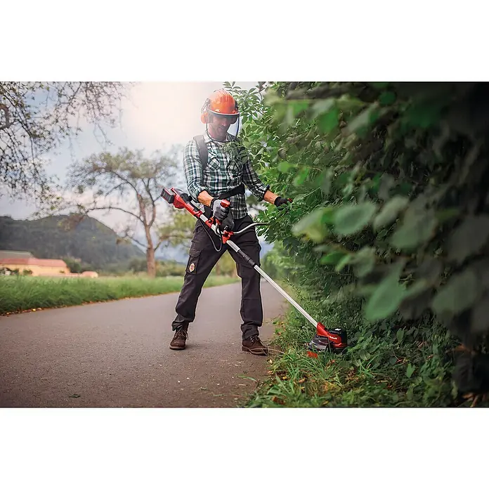 Einhell PXC 18V Desbrozadora de batería Agillo 36/255 - 36 V, 4 Ah, 2 baterías - Hombre con equipo de seguridad recorta arbustos con una desbrozadora roja y negra a lo largo de un camino.
