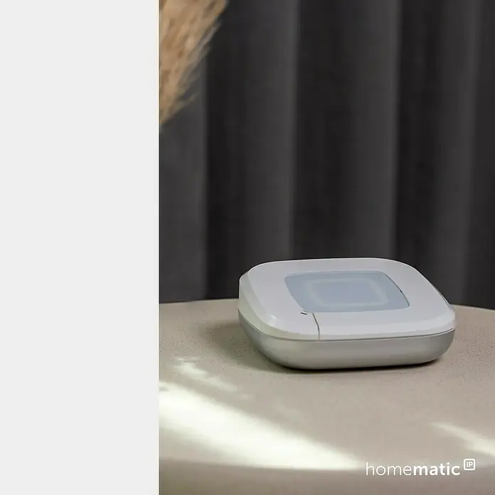 Homematic IP Steuerzentrale Access Point 2 - Weiß, L x B x H: 10,4 x 10,4 x 4 cm - Weiß-silbernes Homematic IP Smart-Home-Gerät mit transluzentem Oberteil, auf einem Tisch vor dunklem Vorhang.