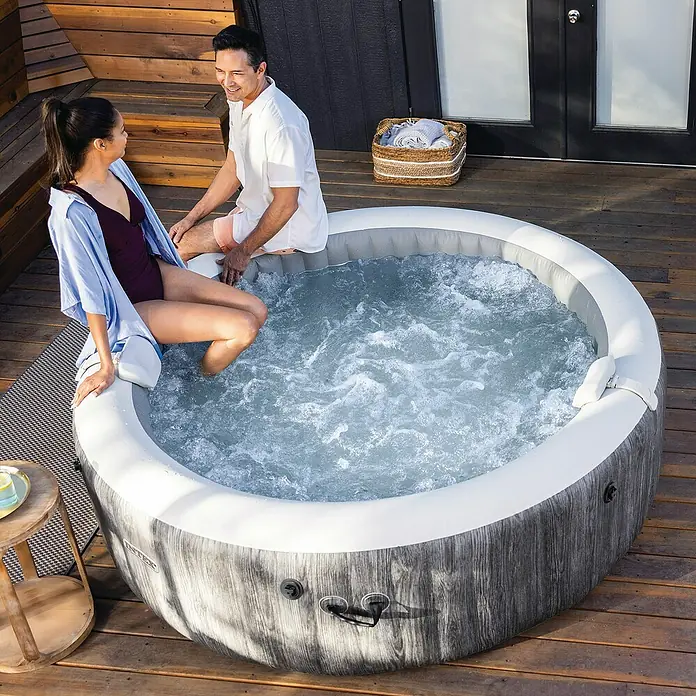 Intex Pure Spa Hidromasaje móvil Greenwood - Ø x Al: 216 x 71 cm, Gris/Blanco, 1.098 l - Dos personas relajándose junto a una bañera de hidromasaje inflable burbujeante con patrón de madera gris en una terraza.