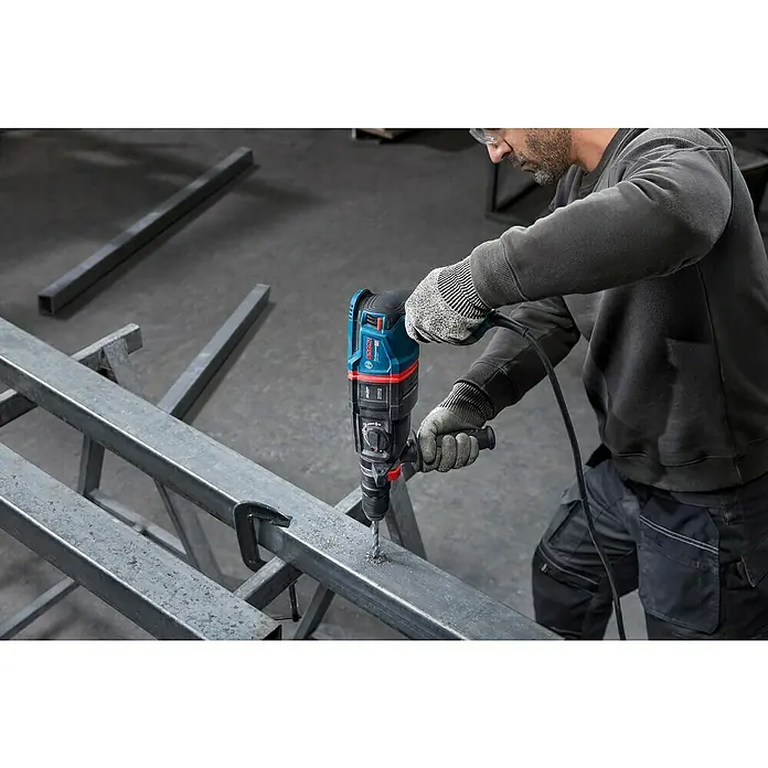 Bosch Professional Bohrhammer-Set GBH 2-26 F - 2,7 J - Eine Person bohrt mit einem blau-schwarzen Bosch Bohrhammer ein Loch in einen Metallträger, mit Handschuhen und Schutzbrille.