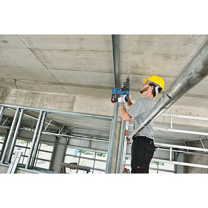 Bosch Professional AMPShare 18V Akku-Kombihammer Pro Heavy Duty GBH 18V-26F - 18 V, Ohne Akku, 2,6 J - Mann mit Schutzhelm und Schutzbrille bohrt mit einem blau-schwarzen Bosch Akku-Bohrhammer in eine Betondecke auf einer Baustelle.
