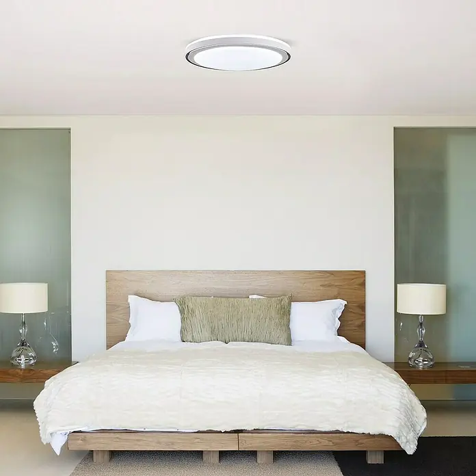 Led Hispania Plafón LED redondo Ítaca - 40 W, Ø x Al: 500 cm x 90 mm, Gris/Plata, Blanco frío - Una moderna lámpara de techo redonda blanca y plateada ilumina un dormitorio luminoso con cama de madera.