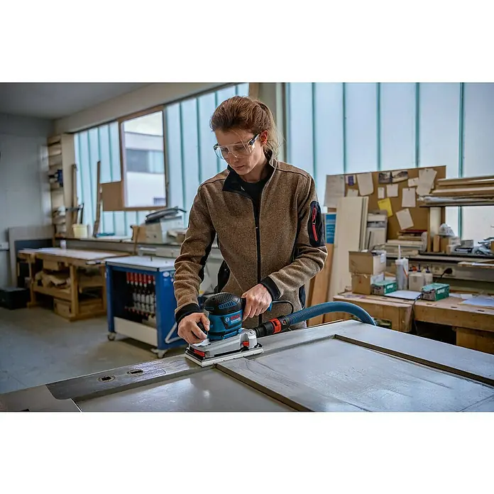 Bosch Professional Elektro-Schwingschleifer GSS 280 AVE - 350 W, 16.000 U/min - 22.000 U/min, Ohne L-Boxx - Frau in Werkstatt schleift graue Holzplatte mit blau-schwarzem Bosch Schwingschleifer.