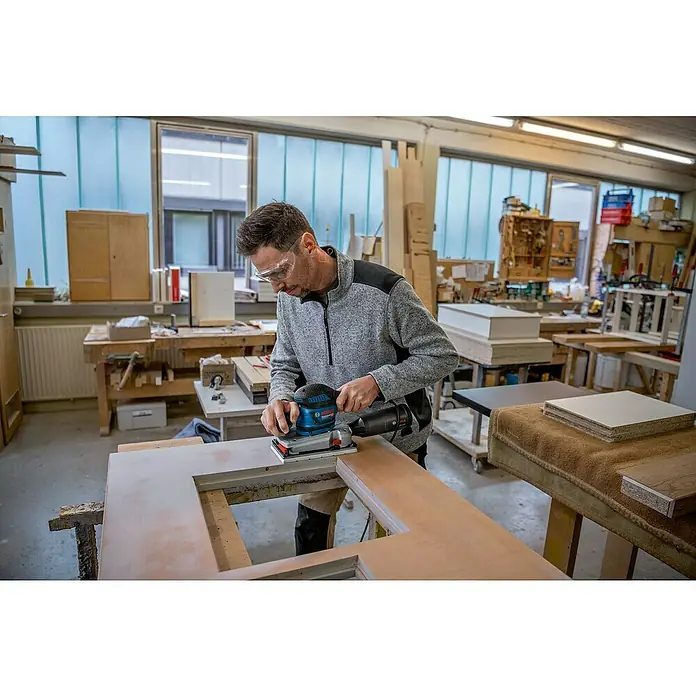 Bosch Professional Elektro-Schwingschleifer GSS 280 AVE - 350 W, 16.000 U/min - 22.000 U/min, Ohne L-Boxx - Mann mit Schutzbrille schleift Holz in einer Werkstatt mit blau-schwarzem Exzenterschleifer.