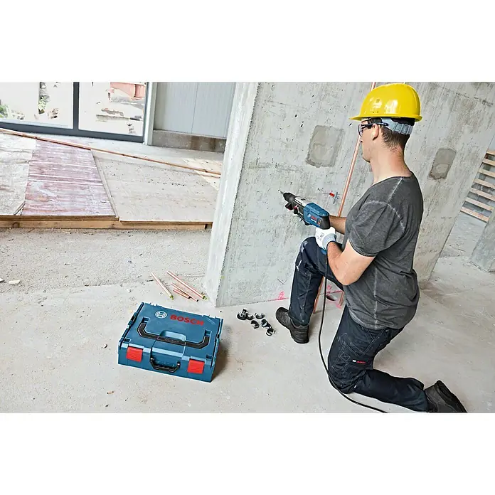 Bosch Professional Bohrhammer GBH 2-28 F - 880 W, 3,2 J, SDS-Plus-Aufnahme - Ein Mann bohrt mit einem blauen Bosch Bohrhammer in eine Betonwand; eine blaue Bosch L-Boxx steht auf dem Boden.