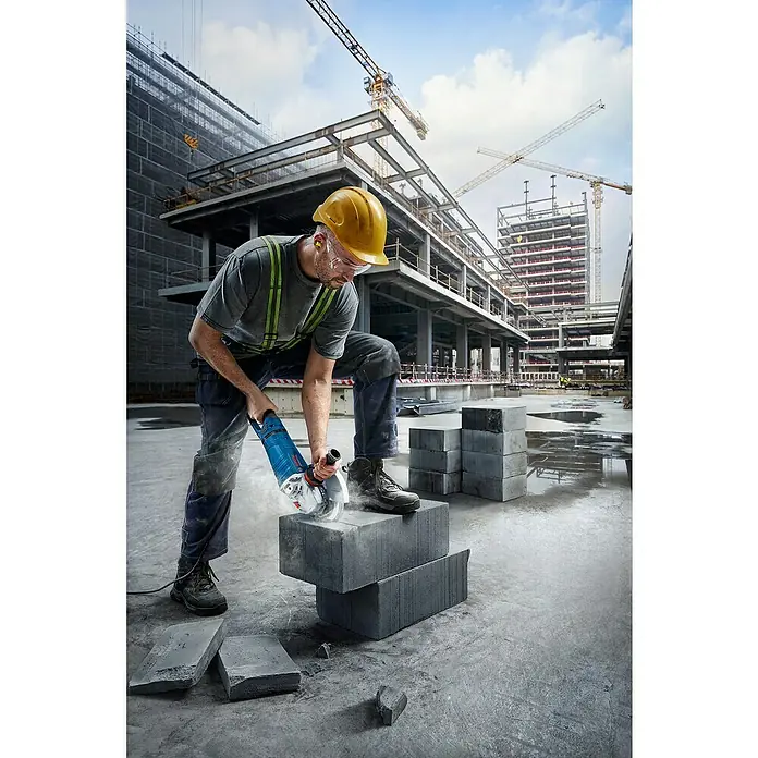 Bosch Professional Elektro-Winkelschleifer GWS 27-230 JR - 2.700 W, Durchmesser Scheibe: 230 mm, Leerlaufdrehzahl: 5.200 U/min - 6.500 U/min - Ein Bauarbeiter schneidet mit einem blauen Bosch Winkelschleifer graue Betonblöcke auf einer Baustelle.