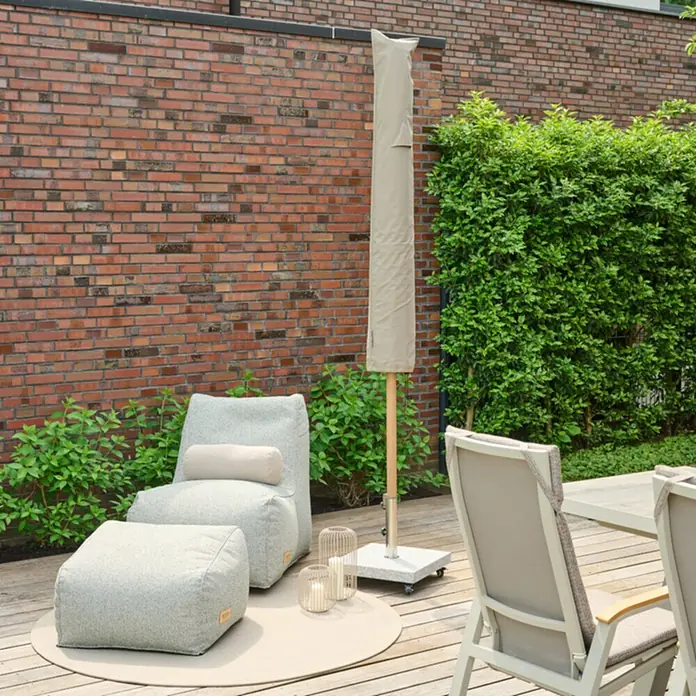 Siena Garden Sonnenschirm Schutzhülle PLUS - Passend für: Schirme mit einem ⌀ bis zu 3,0 m, Polyester, TPU laminiert, Beige - Hellgrauer Outdoor-Sitzsack mit Hocker auf einer Terrasse, daneben ein abgedeckter beiger Sonnenschirm.