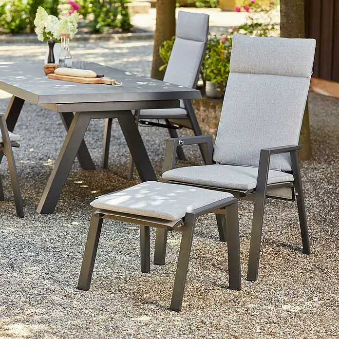 Gartenhocker Valencia - Aluminium, Polypropylen, Ranotex, Anthrazit, B x T x H: 58 x 49 x 46 cm - Dunkelgrauer Outdoor-Sessel mit hellgrauen Polstern und passender Fußstütze, neben einem Tisch auf Kies.