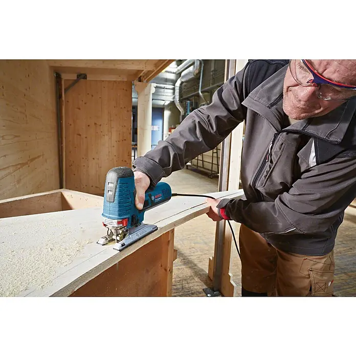 Bosch Professional Elektro-Stichsäge GST 160 CE - 800 W, 800 /min - 3.000 /min - Ein Mann sägt mit einer blauen Bosch Stichsäge präzise ein helles Holzbrett und erzeugt Sägemehl.