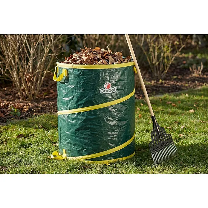 Gardol Pop-Up Gartenabfallsack - 120 l, Höhe: 60 cm, Durchmesser: 50 cm - Großer grüner Gartenabfallsack mit gelben Details, gefüllt mit Blättern, und eine Harke lehnt im Garten daran.