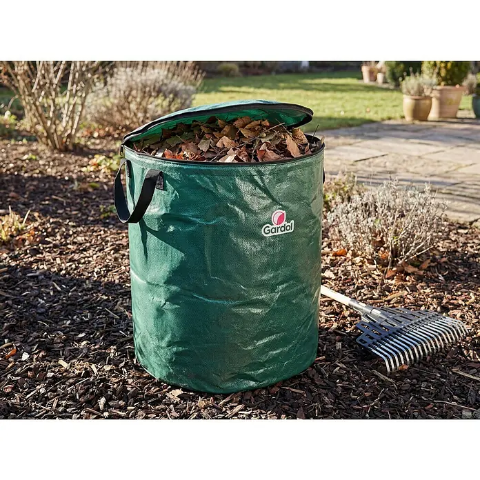 Gardol Comfort Gartenabfallsack - 215 l, Ø x H: 60 x 76 cm - Grüner Gartensack aus Gewebe mit Reißverschlussdeckel, gefüllt mit Herbstlaub, und einem Rechen daneben im Garten.