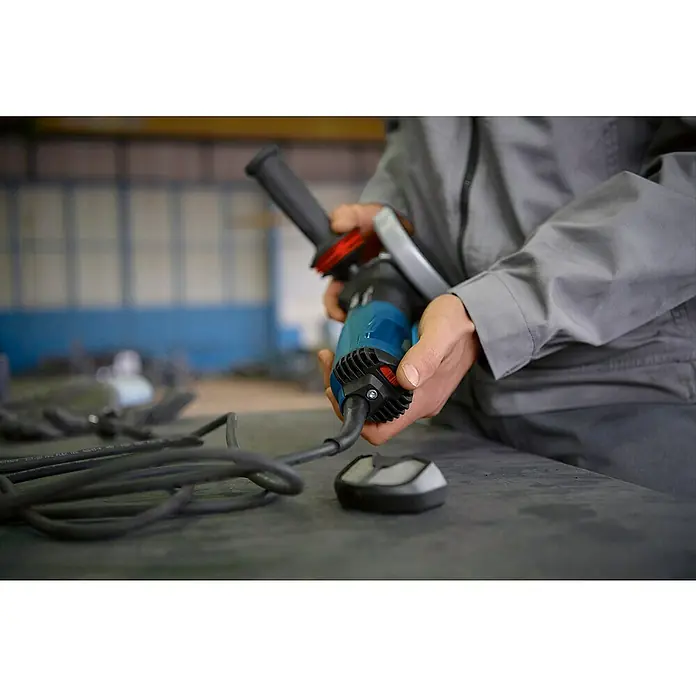 Bosch Professional Elektro-Winkelschleifer PRO GWS 17-125 SB - 1.700 W, Durchmesser Scheibe: 125 mm, Leerlaufdrehzahl: 2.800 U/min - 11.500 U/min - Person hält einen blau-schwarzen Winkelschleifer mit Zusatzgriff und Kabel in einer Werkstatt.