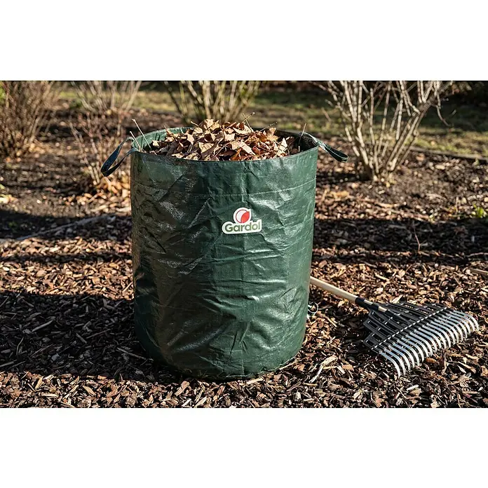 Gardol Gartenabfallsack - 120 l, Höhe: 60 cm, Durchmesser: 50 cm - Ein grüner, robuster Gartenabfallsack, gefüllt mit trockenen Blättern, steht auf Rindenmulch neben einem Rechen.