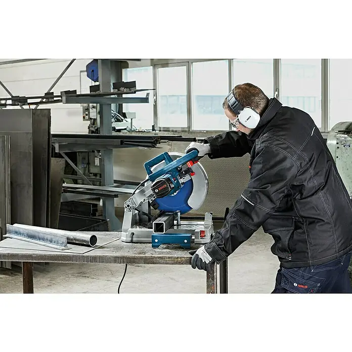Bosch Metallkreissäge GCD 12 JL - 2.000 W, Leerlaufdrehzahl: 1.500 U/min - Ein Mann mit Schutzausrüstung schneidet mit einer blau-silbernen Bosch Trennsäge ein Metallprofil in einer Werkstatt.