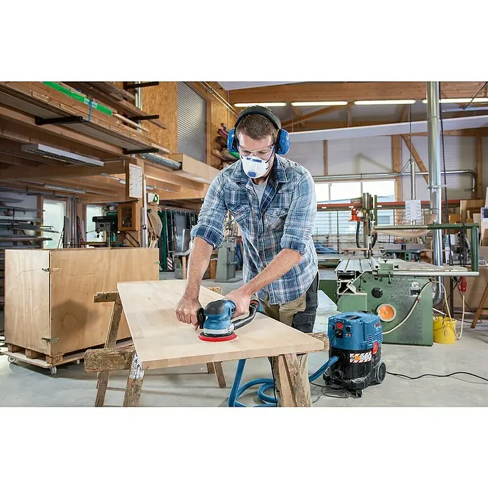 Bosch Professional Elektro-Exzenterschleifer GET 75-150 - 750 W, Durchmesser Schleifteller: 150 mm - Mann schleift ein Holzbrett mit einem blauen Exzenterschleifer, verbunden mit einem Staubsauger, in einer Werkstatt.