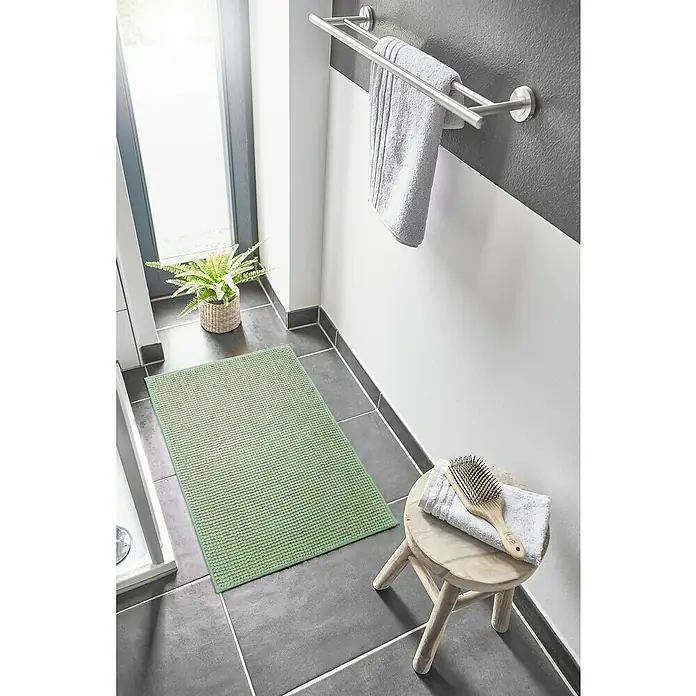 Badteppich Zottel - 50 x 80 cm, Salbei, 100% Polyester - Ein hellgrüner, strukturierter Badteppich liegt auf dunklen Badezimmerfliesen neben einem Duschbereich.