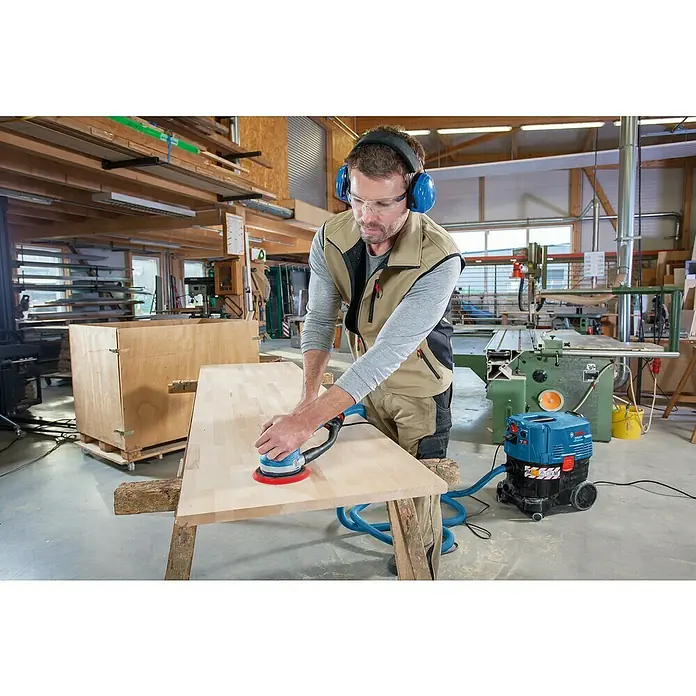 Bosch Professional Elektro-Exzenterschleifer GET 75-150 - 750 W, Durchmesser Schleifteller: 150 mm - Arbeiter schleift Holz mit Exzenterschleifer, verbunden mit Staubsauger, trägt Schutzausrüstung in Werkstatt.