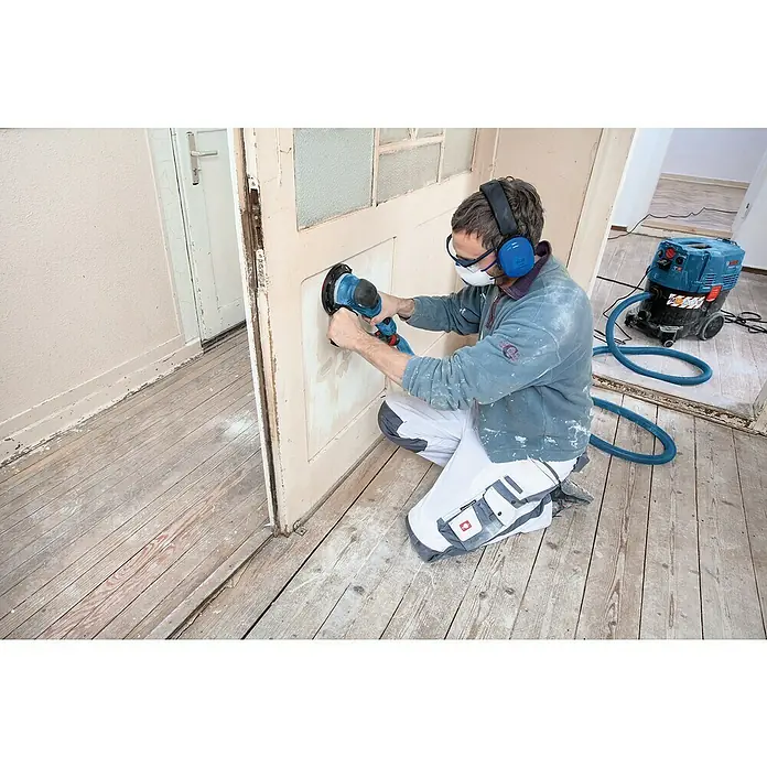 Bosch Professional Elektro-Exzenterschleifer GET 75-150 - 750 W, Durchmesser Schleifteller: 150 mm - Mann schleift Holztür mit blauem Schwingschleifer, an Staubabsauger angeschlossen. Er trägt Schutzausrüstung.