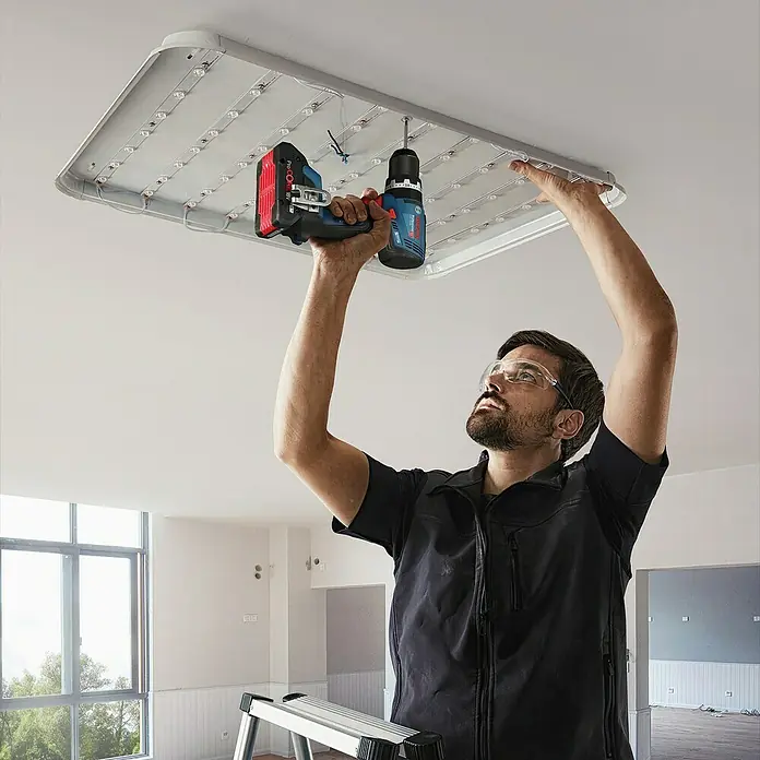 Bosch Professional AMPShare 18V Akku-Schlagbohrschrauber GSB 18V-65 - 18 V, 2 Akkus, 4 Ah, 63 Nm, 2 x 4 Ah Akku, L-BOXX 136 - Mann installiert eine weiße, rechteckige LED-Deckenleuchte an der Decke mit einem blau-schwarzen Akku-Bohrschrauber und roter Batterie.