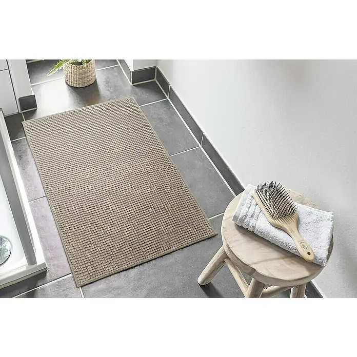 Badteppich Zottel - 50 x 80 cm, Sand, 100% Polyester - Beiger Badteppich mit Noppenstruktur auf dunklem Fliesenboden im Bad, mit Holzhocker, Handtuch und Bürste.