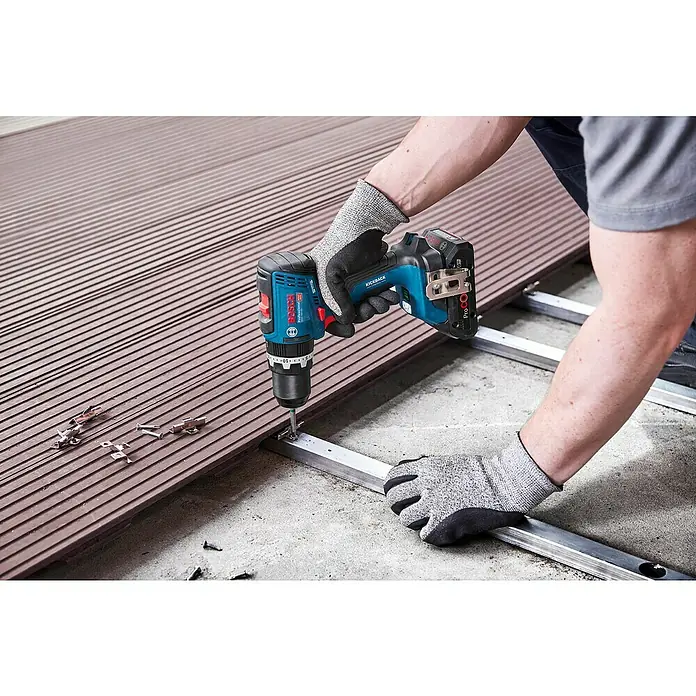 Bosch Professional AMPShare 18V Akku-Schlagbohrschrauber GSB 18V-65 - 18 V, 2 Akkus, 4 Ah, 63 Nm, 2 x 4 Ah Akku, L-BOXX 136 - Nahaufnahme von behandschuhten Händen, die einen blauen Bosch Akku-Bohrschrauber zum Verschrauben einer braunen WPC-Terrassendiele auf Metallunterbau verwenden.
