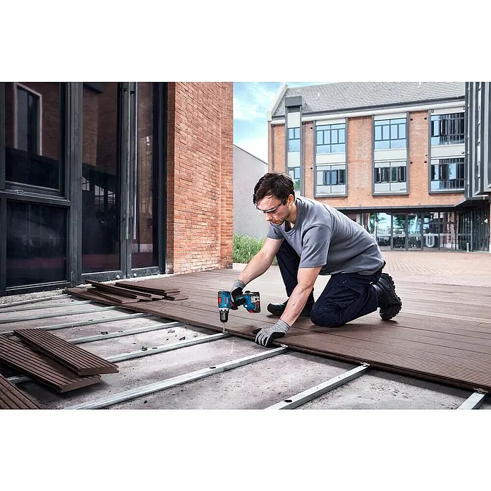 Bosch Professional AMPShare 18V Akku-Bohrschrauber GSR 18V-65 - 18 V, 2 Akkus, 4 Ah, 76 Nm, 2 x 4 Ah Akku, mit L-BOXX-Koffer - Mann mit Schutzbrille und Handschuhen nutzt blauen Bosch Akku-Bohrschrauber zur Installation brauner Terrassendielen auf einer Terrasse.