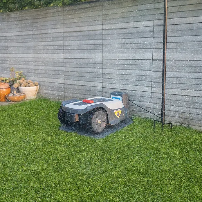 RoboUp Mähroboter T1200 - 1.500 m², Schnittbreite: 20 cm, Kamera-gestützte Navigation, RTK - Ein grau-schwarzer Mähroboter steht auf seiner Matte im leichten Regen, an eine Ladestation angeschlossen.