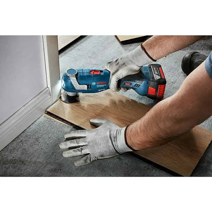 Bosch Professional AMPShare 18V Akku-Multitool GOP 18V-34 - 18 V, Ohne Akku - Bosch Professional 18V Akku-Multifunktionswerkzeug, von behandschuhten Händen gehalten, schneidet Holz nahe einer Fußleiste.