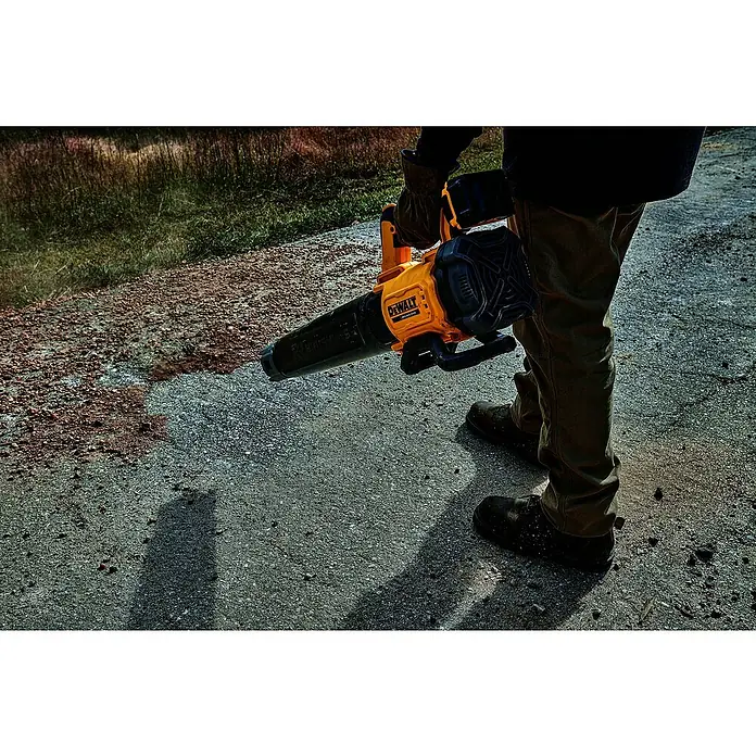 DeWalt XR 18V Akku-Gebläse DCMBL562N-XJ - 18 V, Ohne Akku, Blasgeschwindigkeit: 0 km/h - 200 km/h - Eine Person in Arbeitsstiefeln verwendet einen gelb-schwarzen DeWalt Laubbläser, um kleine Steine und Schmutz von einer grauen gepflasterten Fläche zu entfernen.