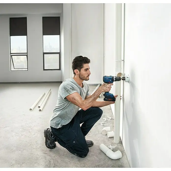 Bosch Professional AMPShare 18V Akku-Schlagbohrschrauber GSB 18V-65 - 18 V, 2 Akkus, 5 Ah, 63 Nm, 2 x 5 Ah Akku, L-BOXX 136N - Mann bohrt mit blauem Akku-Bohrschrauber ein Loch in eine weiße Wand neben weißen Rohren.