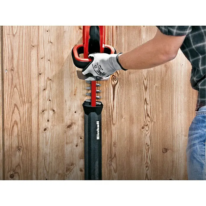 Einhell Power X-Change 18V Akku-Heckenschere GC-CH 1846 Li-Solo - 18 V, Ohne Akku, Schnittlänge: 46 cm - Das Bild zeigt eine Einhell Heckenschere mit Schutzhülle, die von einer Person mit Handschuhen gehalten wird. Der Hintergrund ist eine Holzwand.