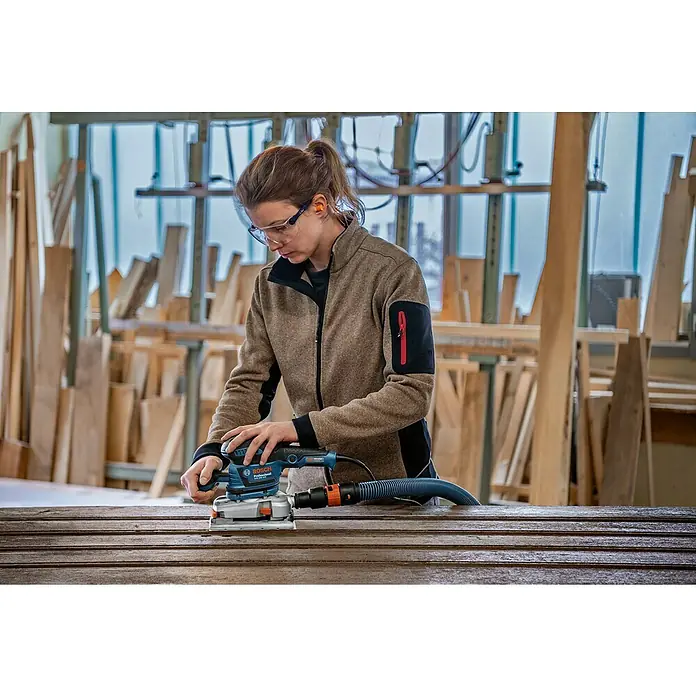 Bosch Professional Elektro-Schwingschleifer GSS 230 AVE - 300 W - Frau schleift Holz mit blau-schwarzem Bosch Schwingschleifer in einer Werkstatt.