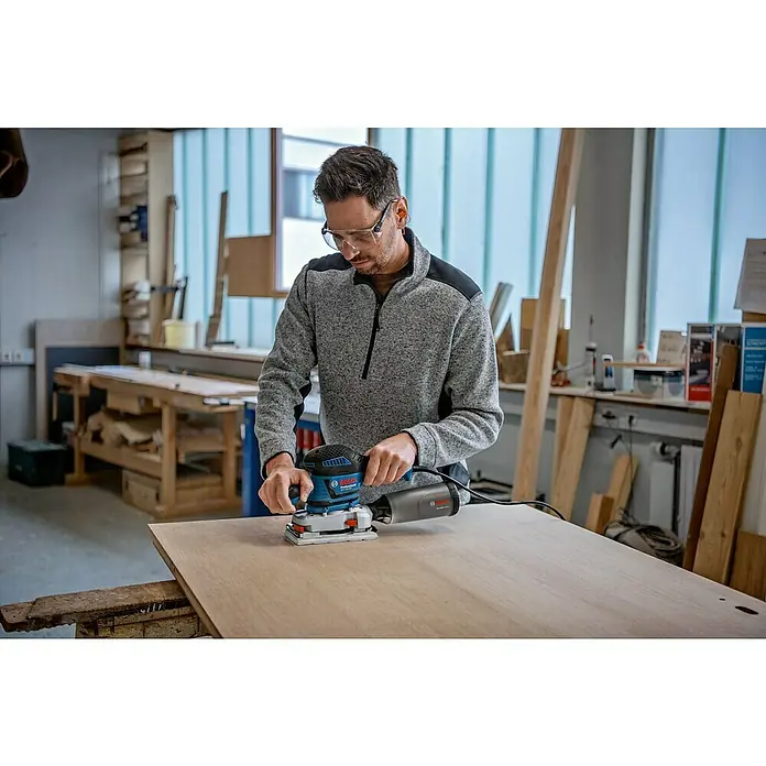 Bosch Professional Elektro-Schwingschleifer GSS 230 AVE - 300 W - Mann mit Schutzbrille schleift eine helle Holzplatte mit einem blauen und schwarzen Bosch Schwingschleifer.