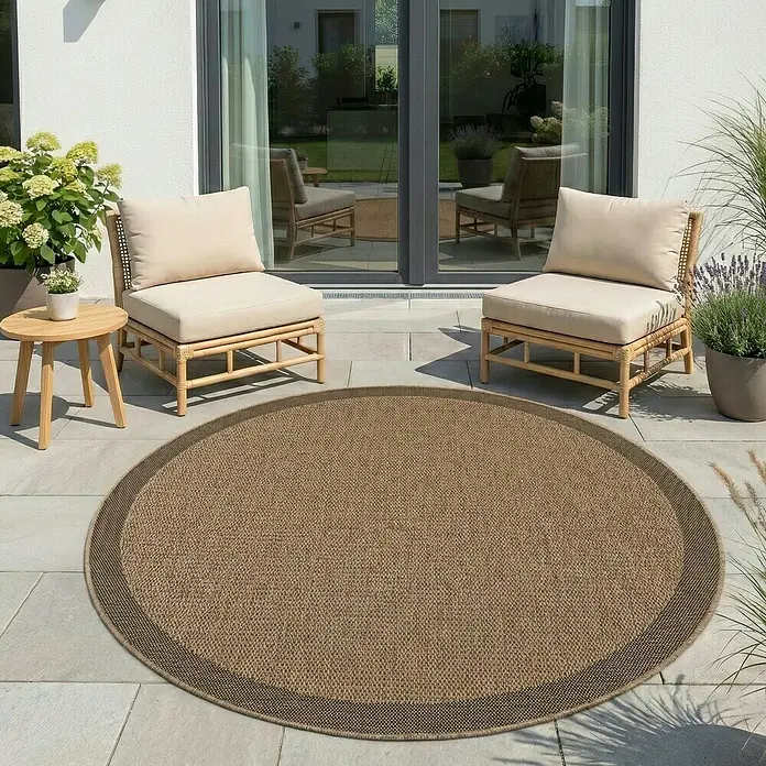 Outdoor-Teppich Timber 1402 - Beige, Durchmesser: 80 cm, 100 % Polypropylen - Runder gewebter Outdoor-Teppich in Beige mit dunklerem braunem Rand, auf einer Terrasse platziert, um einen gemütlichen Sitzbereich zwischen zwei Stühlen zu definieren.