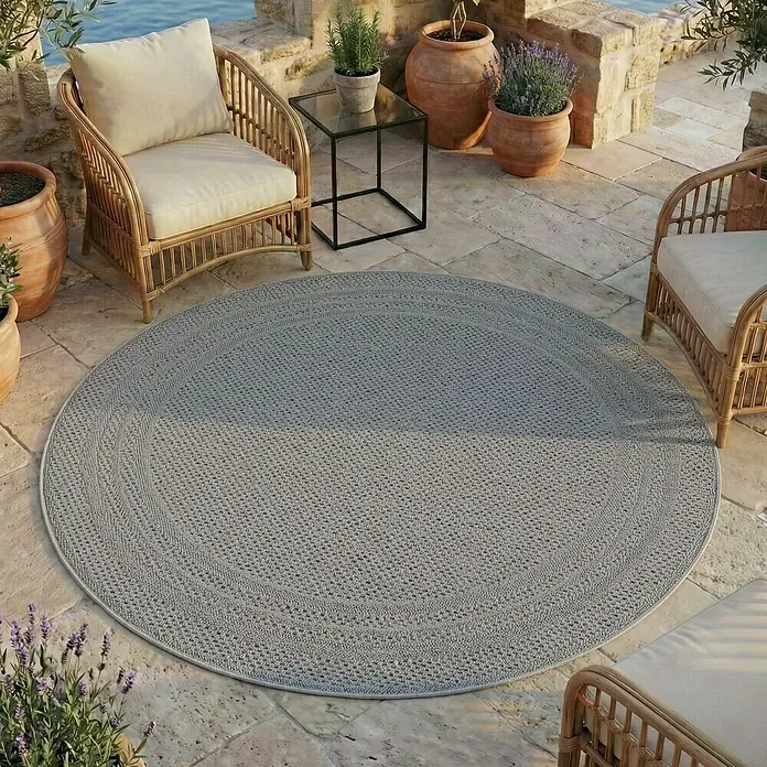 Outdoor-Teppich Desert 1304 - Grau, Durchmesser: 160 cm, 100 % Polypropylen - Ein runder, grauer geflochtener Teppich auf einer Steinterrasse, umgeben von Gartenmöbeln und Topfpflanzen.