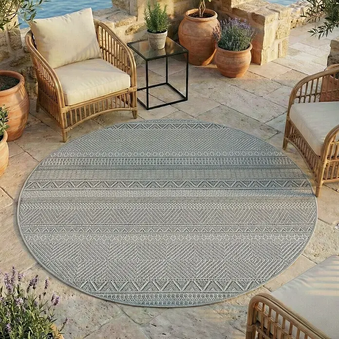 Outdoor-Teppich Desert 1303 - Grau, Durchmesser: 160 cm, 100 % Polypropylen - Ein runder grauer Outdoor-Teppich mit geometrischen Mustern auf einer Steinterrasse inmitten von Gartenmöbeln und Topfpflanzen.