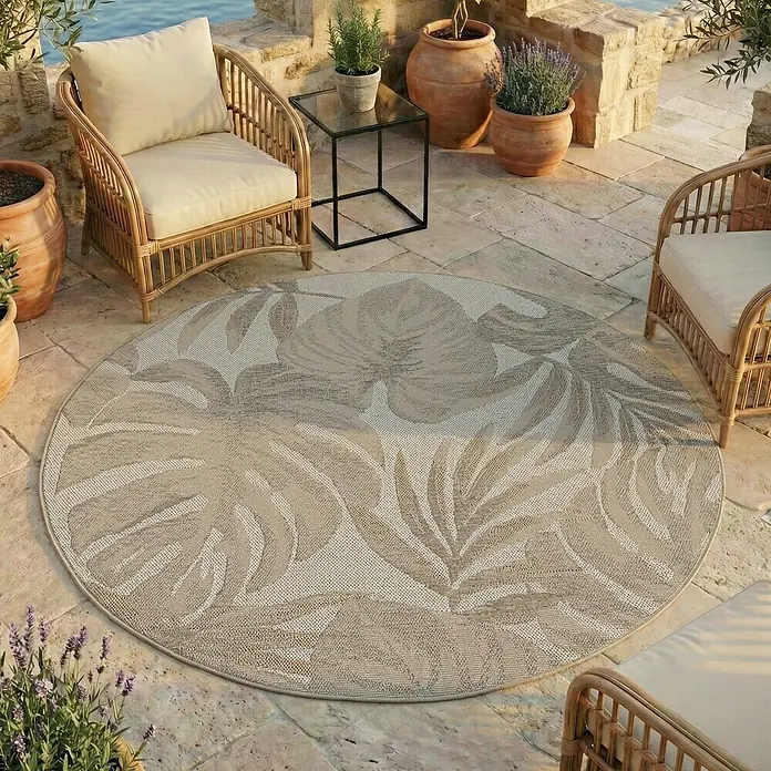 Outdoor-Teppich Desert 1302 - Beige, Durchmesser: 200 cm, 100 % Polypropylen - Ein runder beiger und hellbrauner Outdoor-Teppich mit tropischem Blattmuster auf einer Terrasse mit Rattanmöbeln.