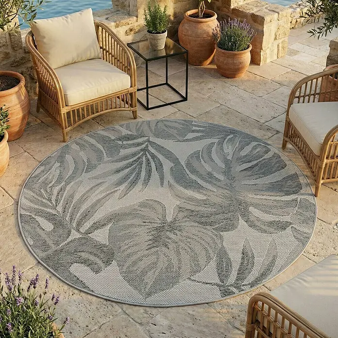 Outdoor-Teppich Desert 1302 - Grau, Durchmesser: 120 cm, 100 % Polypropylen - Runder, gewebter Outdoor-Teppich mit tropischem Blattmuster in Grautönen, auf einer Terrasse.