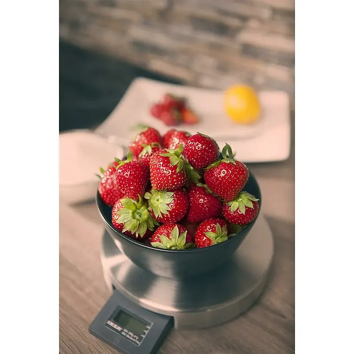Erdbeere Bio - Fragaria ananassa Sweet Mary, Topfgröße: 10 cm - Frische rote Erdbeeren in einer schwarzen Schale auf einer Küchenwaage, mit mehr Obst im unscharfen Hintergrund.