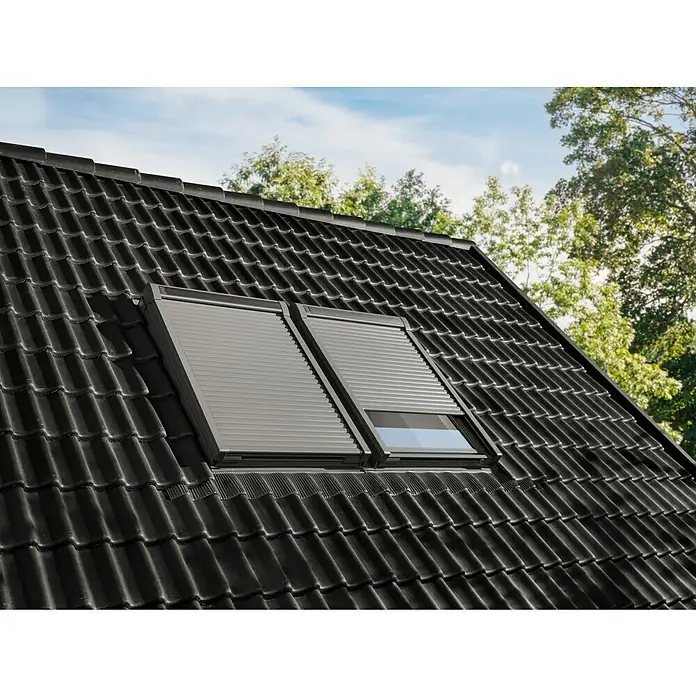 Velux Dachfenster-Rollladen Elektro SML CK06 0000K - Passend für Fenstergröße: CK06, C06, Passend für Fenstertyp: GGL, GGU, GPL, GPU, GHL, GHU, GZL, GLL, GLU, VU und VKU - Zwei graue Außenrollläden auf Dachfenstern, auf dunklem Ziegeldach installiert, einer teilweise geöffnet.