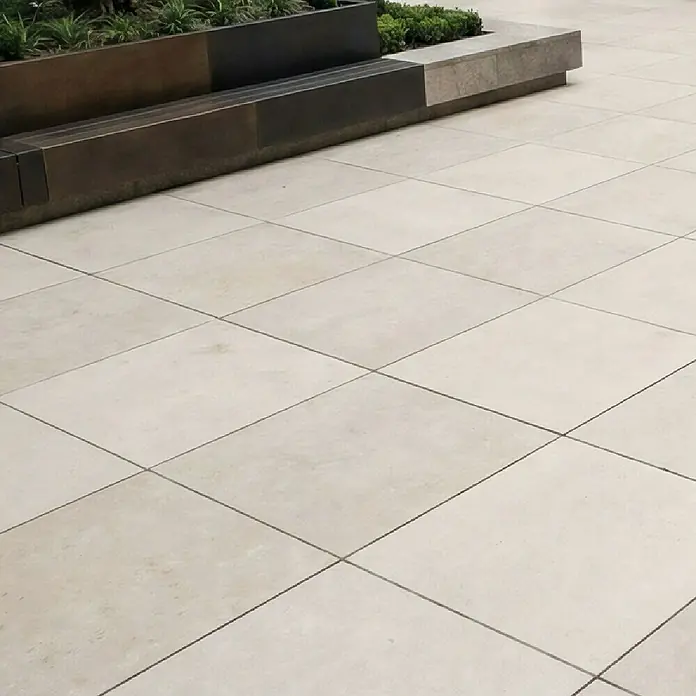 Terrassenfliese Colosseo - 120 x 120 x 2 cm, Pietra, Matt - Grosse, helle Outdoor-Fliesen mit dunklen Fugen, neben einem modernen Pflanzgefäss und einer Bank.