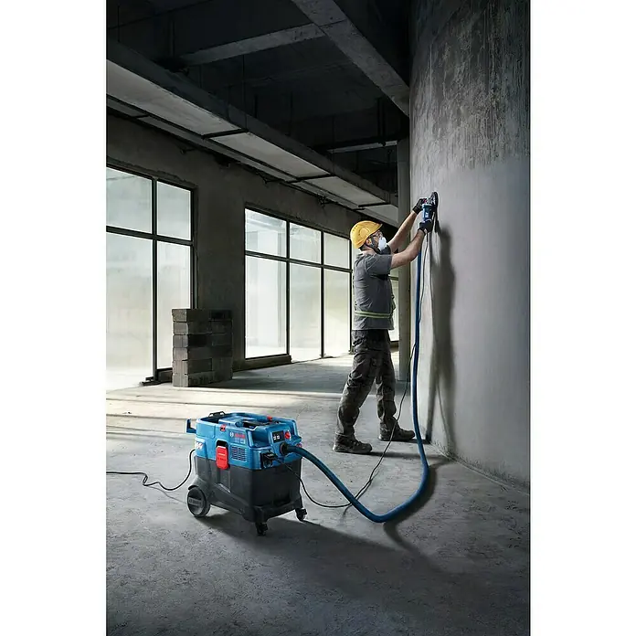Bosch Professional Elektro-Nass- & Trockensauger PRO GAS 400 A - 1.200 W, Behältervolumen: 40 l, Vollautomatisch - Blauer und schwarzer Bosch Industriesauger wird von einem Arbeiter zum Schleifen einer Betonwand mit Staubabsaugung verwendet.